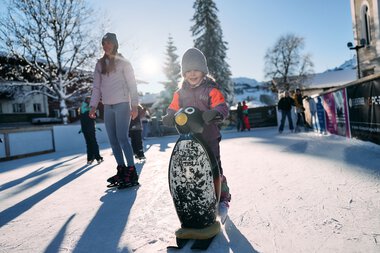 Children ice skating on an ice rink in Riezlern during winter, surrounded by snow-covered scenery and some adults. | © Kleinwalsertal Tourismus eGen • Photographer: Oliver Farys