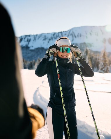 Frau beim Langlaufen im Schnee, trägt eine Skihelm und Skistöcke, vor einer verschneiten Bergkulisse bei Sonnenaufgang oder Sonnenuntergang. | © Kleinwalsertal Tourismus eGen • Fotograf: Oliver Farys