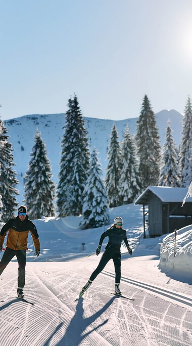 Zwei Skifahrer ziehen ihre Spuren im verschneiten Winterwald beim Langlaufen im Kleinwalsertal, Sonnenschein und klare Sicht. | ©  Kleinwalsertal Tourismus eGen • Fotograf: Oliver Farys
