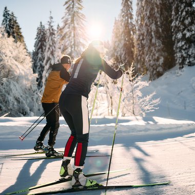 Zwei Skifahrer beim Langlaufen in verschneiter Winterlandschaft, Sonnenlicht durch Bäume, alpine Natur im Hintergrund. | © Kleinwalsertal Tourismus eGen • Fotograf: Oliver Farys