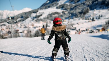 A child skier in winter clothing on a snowy slope, with mountains and other skiers in the background. | © Kleinwalsertal Tourismus eGen • Photographer: Oliver Farys