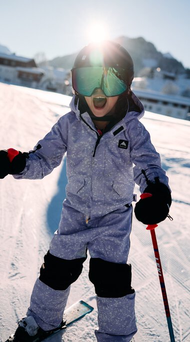 A young child skiing on a slope at a ski resort in Kleinwalsertal, surrounded by mountains, wearing goggles and winter clothing, holding ski poles. | © Kleinwalsertal Tourismus eGen • Photographer: Oliver Farys