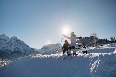 Children skiing on a snow-covered slope in the mountains under sunlight, accompanied by an adult. | © Kleinwalsertal Tourismus eGen • Photographer: Oliver Farys