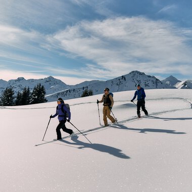 Drei Skitourengeher in schneebedeckter Berglandschaft mit Gipfeln und bewölktem Himmel, bei winterlicher Tour im Kleinwalsertal. | © Kleinwalsertal Tourismus eGen • Fotograf: Oliver Farys