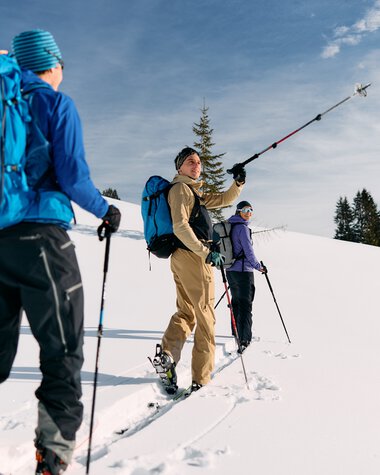 Three skitourers with backpacks in a snowy landscape, one taking a selfie-like photo during a winter tour in Kleinwalsertal. | © Kleinwalsertal Tourismus eGen • Photographer: Oliver Farys