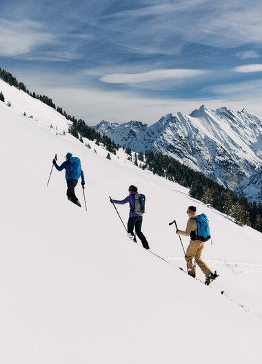 Three backcountry skiers ascending a snowy mountain slope in Kleinwalsertal with mountains in the background. | © Kleinwalsertal Tourismus eGen • Photographer: Oliver Farys