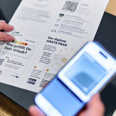 Person holds a digital guest pass and reads a printed document at a table, while someone takes a photo of it with a smartphone. | © Kleinwalsertal Tourismus eGen • Photographer: Oliver Farys