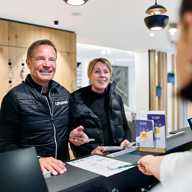 Two smiling people at the reception desk during winter 2025 registration at Kleinwalsertal, served by an employee. | © Kleinwalsertal Tourismus eGen • Photographer: Oliver Farys