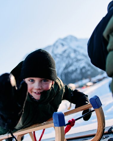 Child sledding in Kleinwalsertal, smiling and wearing winter clothes, with mountains in the background. | © Kleinwalsertal Tourismus eGen • Photographer: Oliver Farys