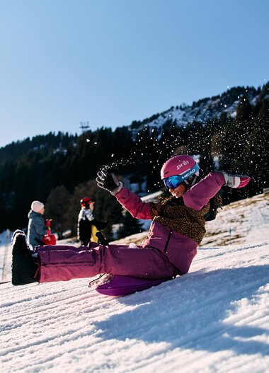 Children sledding in winter at Kleinwalsertal, laughing and playing on a snowy slope surrounded by mountains. | © Kleinwalsertal Tourismus eGen • Photographer: Oliver Farys