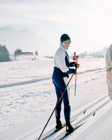 Skifahrer auf der Langlaufloipe im Schnee, umgeben von Bergen und einer Winterlandschaft, mit einer Person im Vordergrund, die einen Skistock hält. | © Kleinwalsertal Tourismus eGen • Fotograf: Oliver Farys