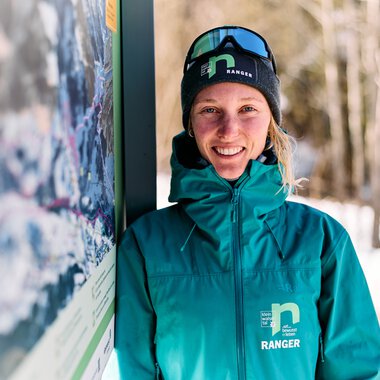A smiling woman in winter gear with ski goggles stands next to an information sign in the snow, with trees and a snowy landscape in the background. | © Kleinwalsertal Tourismus eGen • Photographer: Oliver Farys