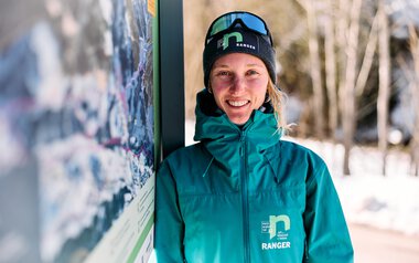 Eine lächelnde Frau in Winterkleidung und mit Skibrille steht neben einem Informationsschild im Schnee, im Hintergrund Bäume und verschneite Landschaft. | © Kleinwalsertal Tourismus eGen • Fotograf: Oliver Farys