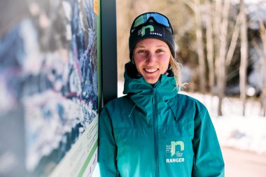 A smiling woman in winter gear with ski goggles stands next to an information sign in the snow, with trees and a snowy landscape in the background. | © Kleinwalsertal Tourismus eGen • Photographer: Oliver Farys