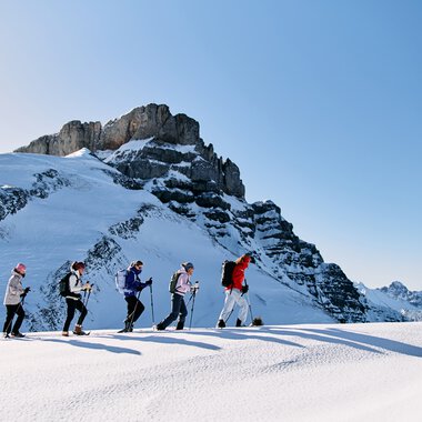 Fünf Leute beim Schneeschuhwandern auf einer verschneiten Berglandschaft im Kleinwalsertal, mit einem hohen Felsmassiv im Hintergrund und klarem blauen Himmel. | © Kleinwalsertal Tourismus eGen • Fotograf: Oliver Farys