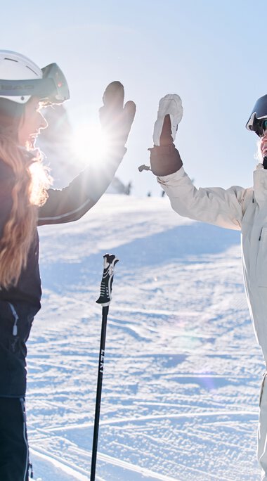 Two female skiers in winter clothing high-fiving on a snowy slope with sunlight in the background at a ski event in Kleinwalsertal. | © Kleinwalsertal Tourismus eGen • Photographer: Oliver Farys