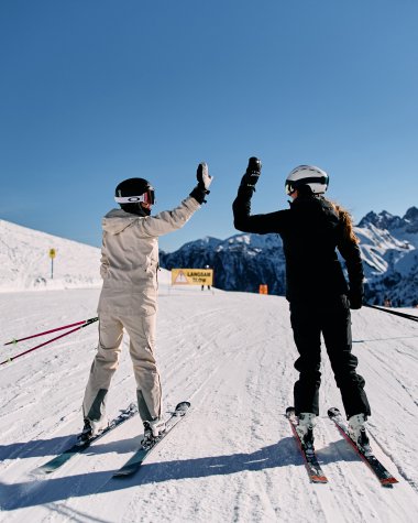 Two skiers giving a high-five on a snowy mountain slope, with the Alps in the background and clear blue sky. | © Kleinwalsertal Tourismus eGen • Photographer: Oliver Farys