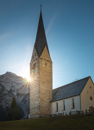 A white church with a stone tower in front of mountain scenery, sunlight behind the tower, blue sky. | © Kleinwalsertal Tourismus eGen • Photographer: Robert Niederwolfsgruber