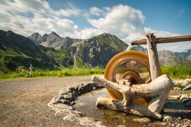 Landschaft mit Bergen und einem alten Wasserrad inmitten einer grünen Wiese, unter einem bewölkten Himmel. | © Kleinwalsertal Tourismus eGen • Fotograf: Robert Niederwolfsgruber