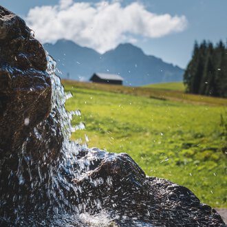 Nahaufnahme eines Wasserfalls oder eines fließenden Bachs in einer grünen, bergigen Landschaft mit Wolken und einem kleinen Haus im Hintergrund. | © Kleinwalsertal Tourismus eGen • Fotograf: Robert Niederwolfsgruber