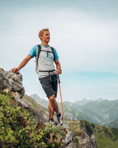 Mann beim Wandern auf einer Steinwand in den Bergen, mit Rucksack und Wanderstöcken, umgeben von grünen Hügeln und einer weiten Berglandschaft. | © Kleinwalsertal Tourismus eGen • Fotograf: Stefan Klauser 