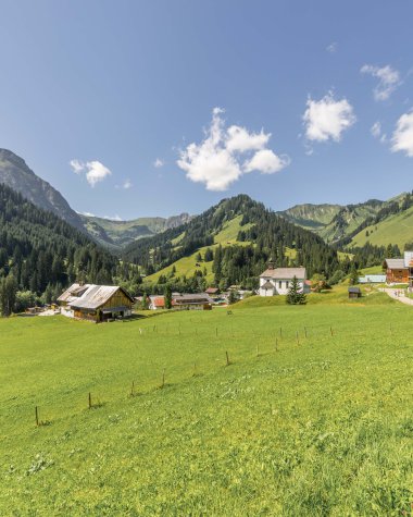 Photo of a green hillside with houses and mountains under a blue sky with clouds. | © Kleinwalsertal Tourismus eGen • Photographer: Steffen Berschin 