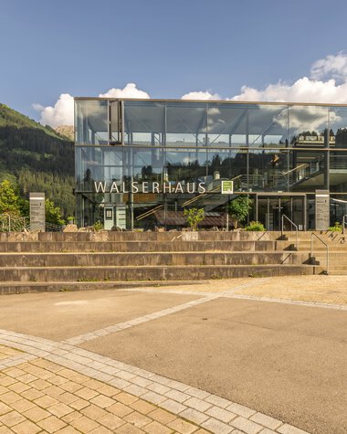 Modern glass building of the Walser House with stairs, surrounded by green mountains and trees on a sunny day. | © Kleinwalsertal Tourismus eGen • Photographer: Steffen Berschin 