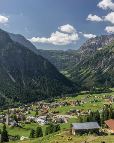 Panoramic view of the village Mittelberg in the Alps, with green hills, mountains in the background, and a blue sky with clouds. | © Kleinwalsertal Tourismus eGen • Photographer: Steffen Berschin 