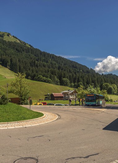 Mountain village with bus parking area surrounded by green hills and forest, sunny summer day, mountain massif in the background. | © Kleinwalsertal Tourismus eGen • Photographer: Steffen Berschin 