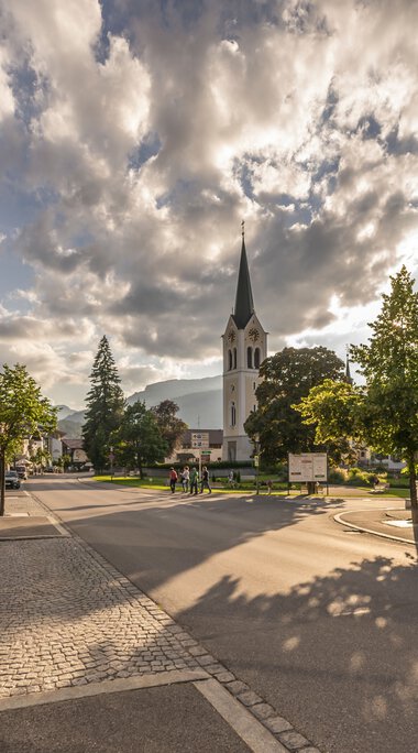 City view with church and sunlight shadows on the street under a cloudy sky in Riezlern. | © Kleinwalsertal Tourismus eGen • Photographer: Stefan Klauser