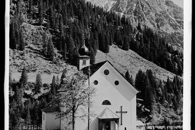 Schwarz-Weiß-Foto einer kleinen Kirche mit Kreuz, umgeben von Bäumen und Bergen, mit Wolfgänger im Hintergrund. | © Kleinwalsertal Tourismus eGen • Karl Max Kessler Archiv
