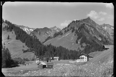 Historisches Schwarz-Weiß-Foto von einem Bergdorf mit mehreren Häusern, umgeben von Wiesen und Wäldern, mit Bergen im Hintergrund. | © Kleinwalsertal Tourismus eGen • Karl Max Kessler Archiv