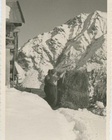 Historical black-and-white photo of a person harvesting hay in a snowy mountain landscape, near a house, during winter. | © Kleinwalsertal Tourismus eGen • Stefan Heim