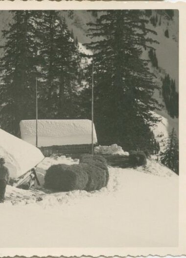 Historical black-and-white photo of a house in snowy landscape, surrounded by pine trees, with a person standing outside. | © Kleinwalsertal Tourismus eGen • Stefan Heim