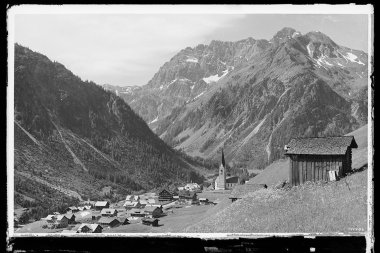 Historisches Schwarz-Weiß-Foto des Ortes Mittelberg im Kleinwalsertal, umgeben von Bergen, mit Häusern und einer Kirche im Zentrum. | © Kleinwalsertal Tourismus eGen • Karl Max Kessler Archiv