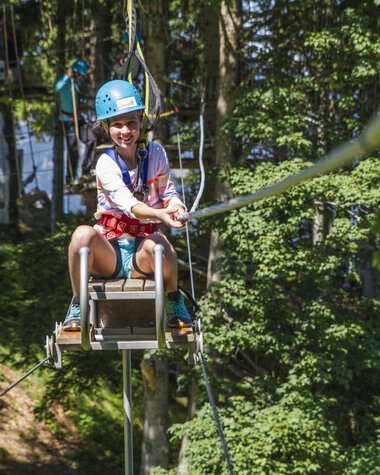 Junge Frau beim Klettergarten am Söllereck, auf einer Seilbahn mit Helm lachend. | © Oberstdorf · Kleinwalsertal Bergbahnen