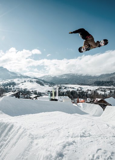Snowboarder airborne over a jump ramp in a snowy winter landscape under sunlight. | © Kleinwalsertal Tourismus eGen • Crystal Ground • Photographer: Stefan Eigner