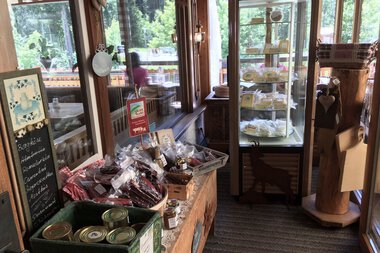 Innenansicht eines kleinen Hofladens mit Produkten auf einem Holzregal und in einem Kühlschrank, Flügeltüren im Hintergrund, viel Naturlicht. | © Kleinwalsertal Tourismus eGen • Walser Buura Café Baad Grund