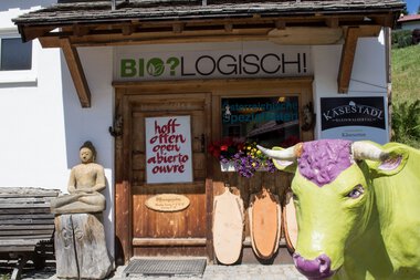 Eingang zum Kasestadl Hofladen in Hirschegg, mit einem begrünten Dach, einem Holzschild, einem Butterschaf-Figur und einem Sitzbank, im Hintergrund blauer Himmel. | © Kleinwalsertal Tourismus eGen • Käsestadl