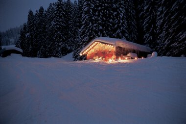 A snow-covered wooden house decorated with Christmas lights in a winter landscape surrounded by tall pine trees. | © Kleinwalsertal Tourismus eGen • Max Hütte • Photographer: Hans Wiesenhofer