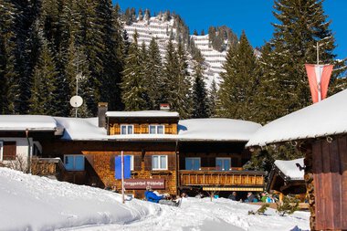 Snow-covered chalets in a mountain village during winter, with pine trees and a steep snowy slope in the background. | © Kleinwalsertal Tourismus eGen • Bühlalpe • Photographer: Frank Drechsel