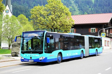 A blue bus is parked at a stop in a rural area, surrounded by trees and houses. | © Kleinwalsertal Tourismus eGen