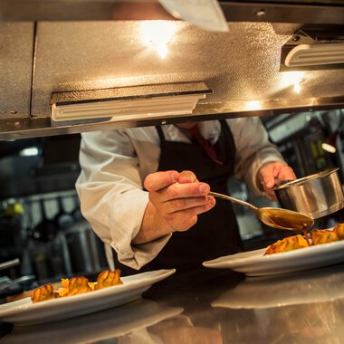 Ein Koch serviert sorgfältig eine Sauce auf vorgerichtete Gerichte in einer professionellen Küche. | © Kleinwalsertal Tourismus eGen • Fotograf: Oliver Farys