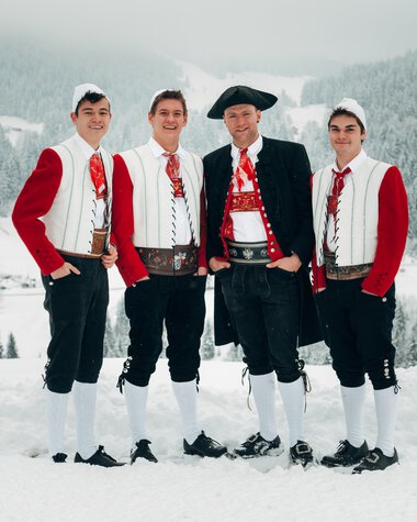 Vier junge Männer in Tracht vor verschneiter Berglandschaft, lachen und posieren gemeinsam im Winter. | © Kleinwalsertal Tourismus eGen • Fotograf: Stefan Klauser