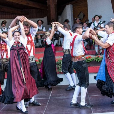 Dance group in traditional costumes performing at a folk festival in Kleinwalsertal, with musicians in the background. | © Kleinwalsertal Tourismus eGen • Photographer: Frank Drechsel