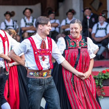 Tanzpaar in traditionellen Alpinskostümen bei einem Alphornfestival in Kleinwalsertal, alle umgeben von weiteren Tanzenden in ähnlicher Tracht. | © Kleinwalsertal Tourismus eGen • Fotograf: Frank Drechsel