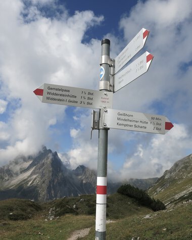 Wanderwegweiser in einer Berglandschaft mit Gipfeln und Hügeln, bei bewölktem Himmel. | © Kleinwalsertal Tourismus eGen • Fotograf: Antje Pabst