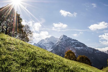 Landscape scene with green grass in the foreground, mountains in the background, and sunlight shining from the top left corner. | © Kleinwalsertal Tourismus eGen