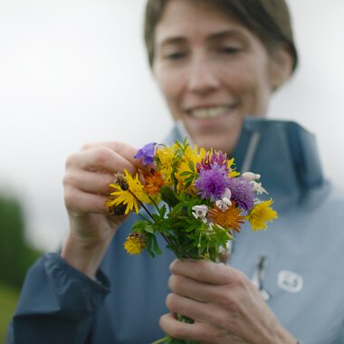 Frau beim Pflücken eines bunten Blumenstraußes im Freien, mit einem Lächeln auf dem Gesicht. | © Kleinwalsertal Tourismus eGen • Fotograf: Clemens Paul