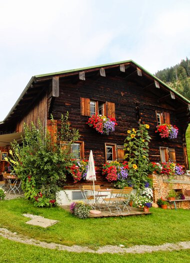 Rustikale, holzgetäfelte Alpenhütte mit bunten Blumen an den Fensterkästen, umgeben von Grünland und mit Bergsicht im Hintergrund. | © Ralf Malzer